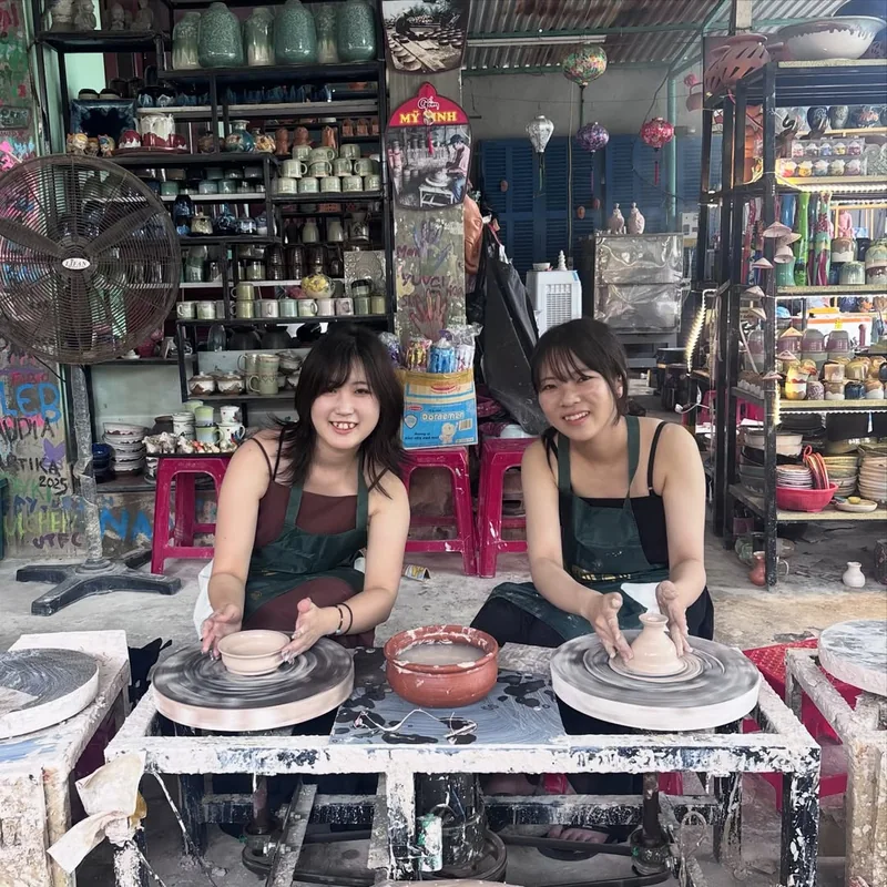 Two visitors smiling at Thanh Ha pottery workshop in Hoi An — pottery wheels and ceramic products displayed throughout the studio
